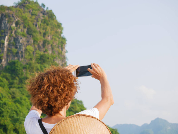 Femme avec chapeau traditionnel utilisant le téléphone à Halong Bay, Vietnam. Tourisme voyageant en croisière parmi les pinacles rocheux de Ha Long Bay dans la mer. Dame caucasienne s'amuser en vacances à célèbre point de repère. - Photo, image