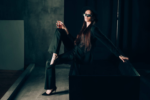 girl in a black trouser suit and hat posing near a black bath. A glass with alcohol in hand. Stylish sunglasses - Photo, Image