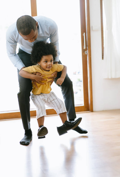 Een gelukkige Afro-Amerikaanse vader draagt een zoontje het huis in terwijl het kind aan het spelen was op zijn vaders schoenen na het werk. Welkom terug thuis papa of familie reünie concept. - Foto, afbeelding