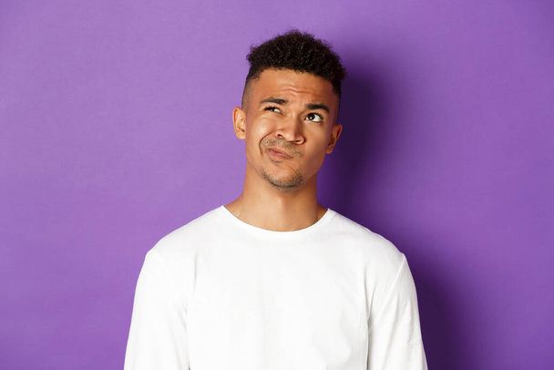 Close-up of indecisive african-american man making choice, looking perplexed at upper left corner, thinking about something complicated, standing over purple background - Foto, Bild