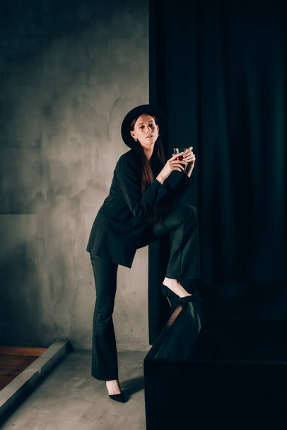 girl in a black trouser suit and hat posing near a black bath. A glass with alcohol in hand. Stylish sunglasses - Foto, imagen