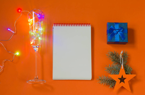 New Year concept. A notebook for writing down goals or plans for the coming year. Christmas toys, blue gift box, garland on an orange background. Flat lay, top view, lifestyle. Copy space - 写真・画像