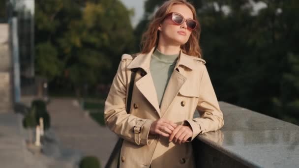 Krásná stylová dívka nosí sluneční brýle a trenč kabát těší slunečné počasí během procházky ulicí města - Záběry, video
