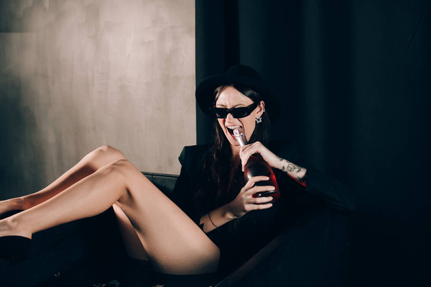 girl in a black jacket and hat posing in a black bath. A bottle of rose champagne wine in hand. Stylish sunglasses. Long naked sexy legs - Foto, immagini