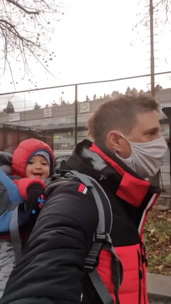 Egy apa, aki orvosi védőmaszkot visel egy gyerekkel a parkban egy hordozóban. - Felvétel, videó