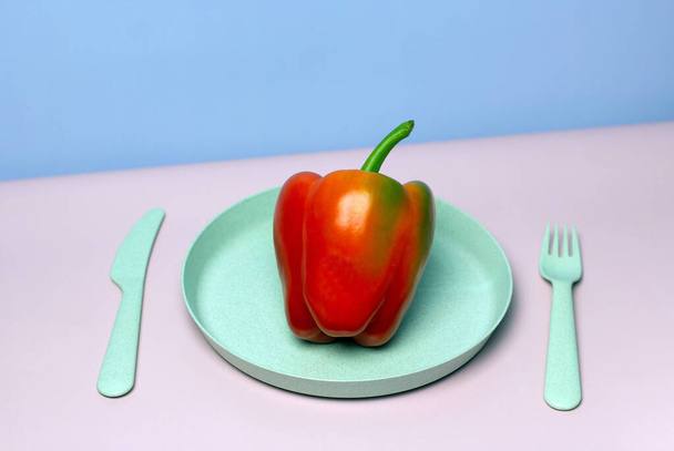 Mise en page tendance avec du poivron rouge sur une assiette avec une fourchette et un couteau. Fond rose et bleu. Concept de nutrition saine. Concept de nourriture minimale. Copier la carte papier espace. - Photo, image