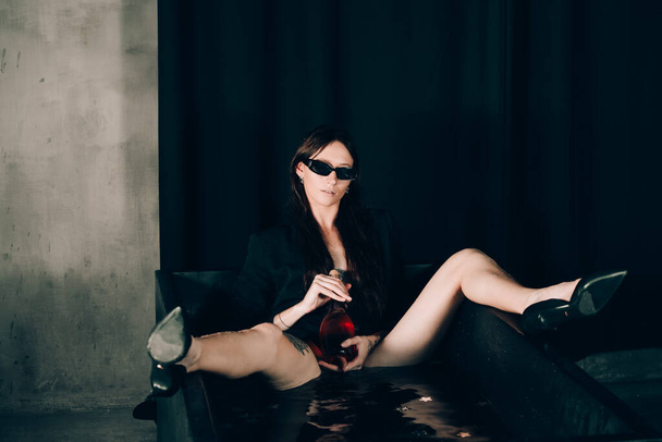 Ένα κορίτσι με μαύρο μπουφάν σε ένα μαύρο μπάνιο. Ανοίγω ένα μπουκάλι κρασί με σαμπάνια τριαντάφυλλο. Κομψά γυαλιά ηλίου. Μακριά γυμνά σέξι πόδια - Φωτογραφία, εικόνα