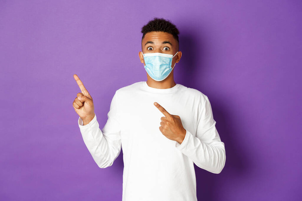 Fogalom a koronavírus, a karantén és az életmód. Lenyűgözött afro-amerikai férfi orvosi maszkban, hirdetést mutat, a bal felső sarokra mutat és mosolyog, a lila háttér fölött áll - Fotó, kép