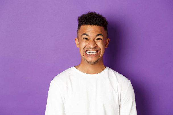 Close-up of funny african-american guy in white sweatshirt, clenching teeth and grimacing at camera, pulling a forced smile, standing over purple background - Foto, immagini