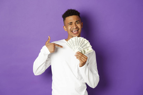 Immagine di sfacciato ragazzo afro-americano che sorride, si vanta con una grande somma di denaro, punta al denaro, in piedi su sfondo viola - Foto, immagini
