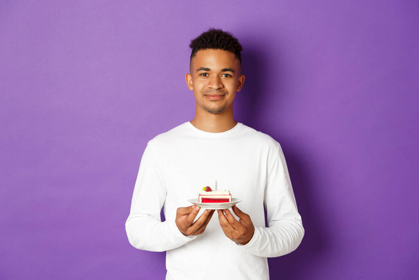Smiling african-american man celebrating his birthday, holding b-day cake with candle, standing over purple background - Photo, image
