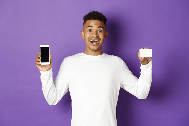 Portrait of handsome african-american man, looking amazed, showing smartphone screen and credit card, standing over purple background - Photo, Image