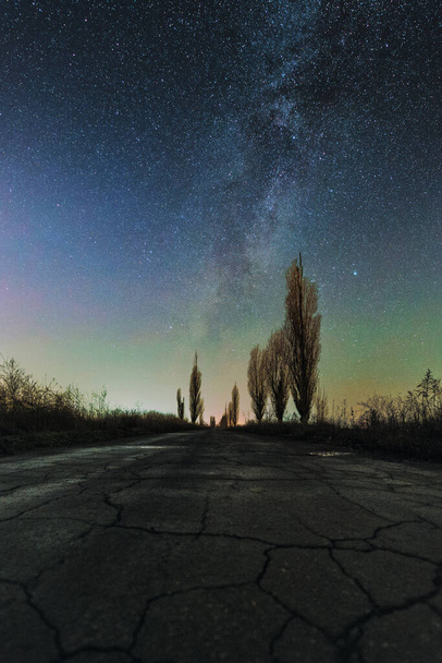 Paisagem astronômica de estrada quebrada e nebulosas da Via Láctea sobre ela. Árvores sem folhas de inverno. À noite. - Foto, Imagem
