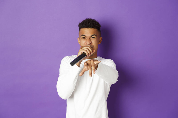 Kép a pimasz afro-amerikai srác énekel mikrofonban, rappel és mutatva kamera, álló felett lila háttér - Fotó, kép