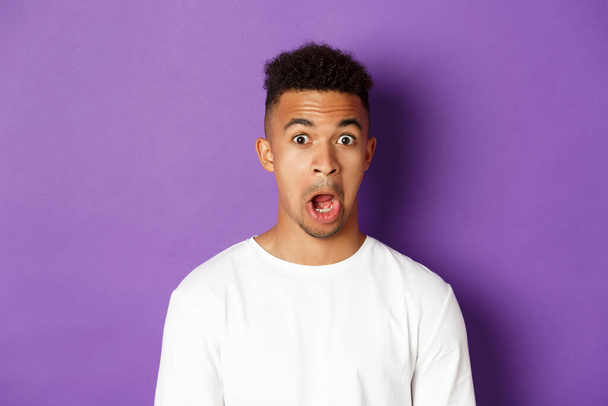 Close-up of impressed african-american man, drop jaw and stare at something amazed, standing over purple background - Foto, immagini