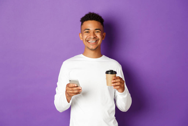 Porträt eines gutaussehenden afrikanisch-amerikanischen Typen, der zufrieden lächelt, Kaffee trinkt und mit dem Handy im Netz surft, vor lila Hintergrund - Foto, Bild