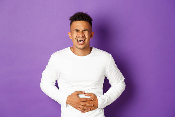 Image of african-american man in white sweatshirt, touching belly and screaming in pain, having stomach ache, food poisoned standing over purple background - Photo, Image