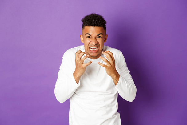 Image of angry african-american young man, shouting and clenching fists from hatred, looking mad and pissed-off at camera, standing over purple background - Photo, Image