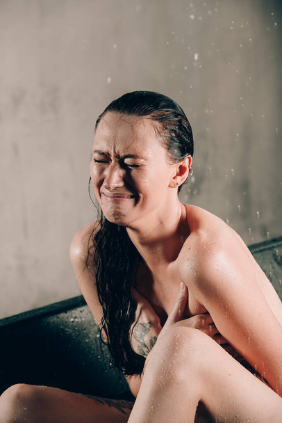 Fiatal, csinos barna nő tetoválásokkal a fürdőben, vízzel és virágokkal. Meztelen modell egy szokatlan fotózáson sírva egy fekete fürdőben. - Fotó, kép