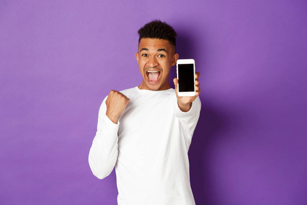 Emocionado hombre afroamericano mostrando la pantalla del teléfono móvil, ganando algo en línea, regocijándose por el logro de la aplicación, de pie sobre el fondo púrpura - Foto, Imagen