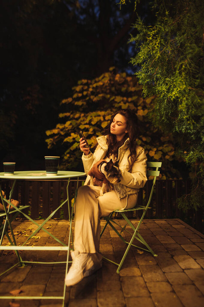 カジュアルな服装の美しい女性は、彼女の腕の中に犬と一緒にカフェでテラスのテーブルに夕方に座って、スマートフォンを使用しています。垂直. - 写真・画像