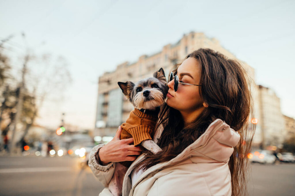 Portrét stylové ženy stojící se psem v náručí na pozadí městského panoramatu a dívající se stranou. Procházka se svým domácím mazlíčkem ve večerním městě. - Fotografie, Obrázek