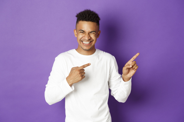 Зображення красивого і безтурботного афроамериканця в білому светрі, вказуючи пальці праворуч і сміючись над чимось смішним, що стоїть над фіолетовим тлом
 - Фото, зображення