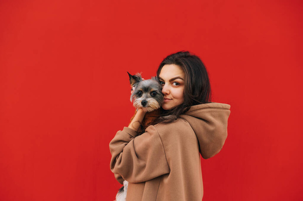 Retrato de uma mulher bonita com um pequeno cão em seus braços isolado em um fundo vermelho, olhando para a câmera e sorrindo. Espaço de cópia - Foto, Imagem