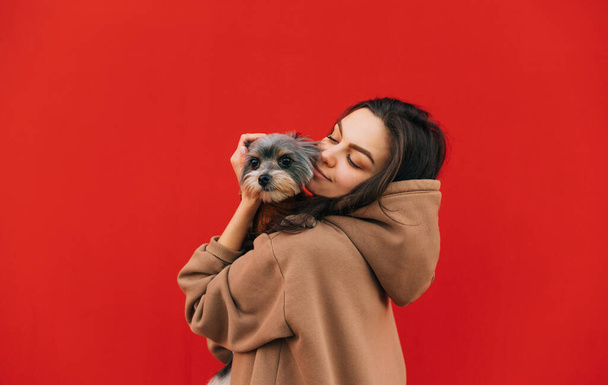 Mujer sonriente de pie sobre un fondo de una pared roja con un perrito lindo en sus brazos, abrazando a una mascota. Señora lleva una mascota en sus brazos, aislada. - Foto, Imagen