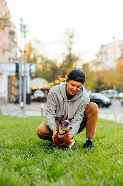 Guy σε casual ρούχα κάθεται στο γκαζόν με φόντο την πόλη με ένα σκυλί biewer terrier σε ένα λουρί και παίζει με ένα χαμόγελο στο πρόσωπό του. Αναψυχή με ένα κατοικίδιο ζώο. - Φωτογραφία, εικόνα
