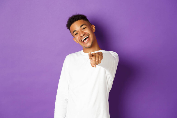 Kuva komea nuori afrikkalais-amerikkalainen mies valkoisessa paidassa, osoittaa sormella kameraa ja hymyilee, valitsee sinut, seisoo purppurainen tausta - Valokuva, kuva