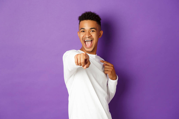 Portrait d'homme afro-américain joyeux en sweat-shirt blanc, vous choisissant, pointant du doigt la caméra pour féliciter avec quelque chose, debout sur fond violet - Photo, image