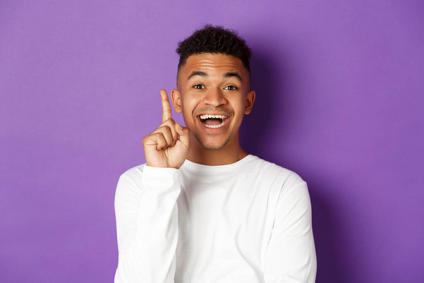 Afbeelding van doordachte Afrikaans-Amerikaanse man hebben een idee, het opsteken van de vinger en geven suggestie, staan in witte sweater over paarse achtergrond - Foto, afbeelding