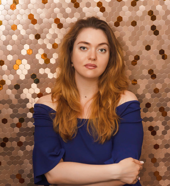 One 30-year-old European woman in a blue dress on a gold glittering background. Long thick reddish hair. Brunette. Blue dress. Calm expression. Confidence. Self-assessment. Beautiful girl. - Photo, image