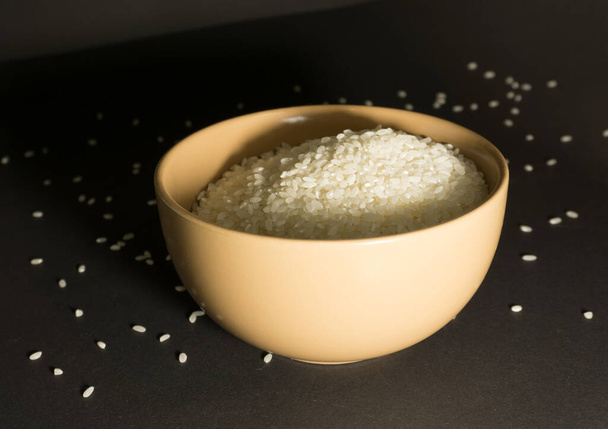 Witte ongekookte rijst in een kom met verspreide korrels in de buurt op een zwarte achtergrond - Foto, afbeelding