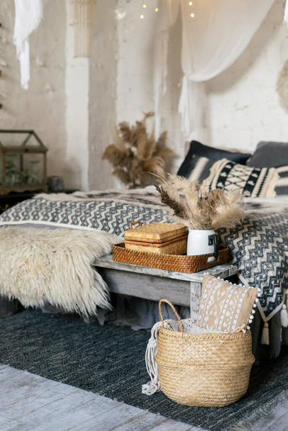 Still life concept. Vertical photo of cozy apartment in boho style interior with comfort bedroom, plaid, pillows and blanket on bed, wooden bench seat, dry plants in vase, home decor in wicker basket - Foto, Imagen