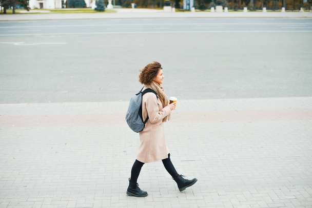 Genç bir kadın elinde bir bardak sıcak içecekle şehirde yürüyor. - Fotoğraf, Görsel