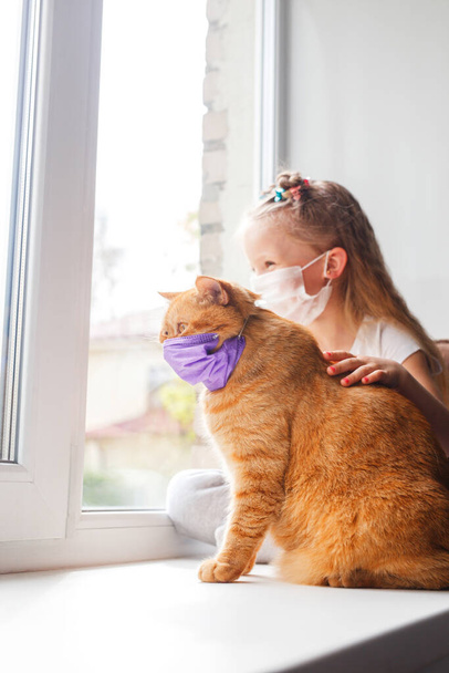 Little girl and cat a in protective masks sitting on window at home - Fotoğraf, Görsel