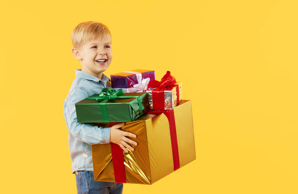 Portrait of happy cute child boy holding gift boxes and looking at camera isolated on yellow background - Fotoğraf, Görsel