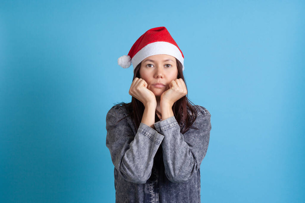 mock up of serious, sad Asian young woman in Santa Claus hat resting her head on her clenched hands on a blue background - Фото, изображение