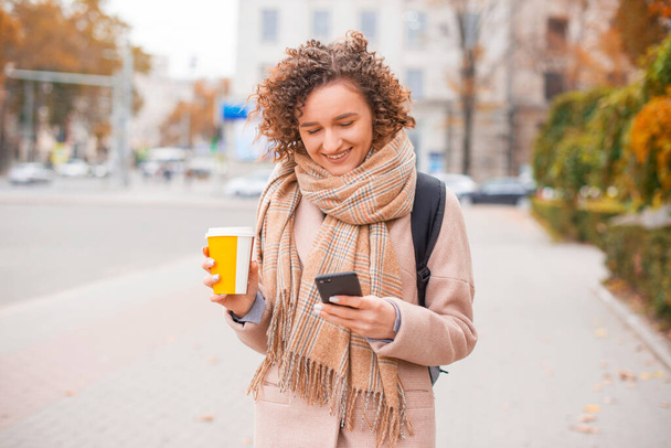 Nainen kävelee ja pitää kupin kuumaa juomaa tekstailee ja hymyilee puhelimessaan kaupungissa. - Valokuva, kuva