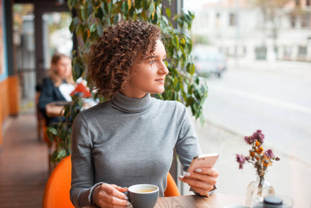 Μια νεαρή γυναίκα κάθεται σε ένα τραπέζι με ένα φλιτζάνι καφέ και ένα τηλέφωνο είναι lookig μακριά - Φωτογραφία, εικόνα