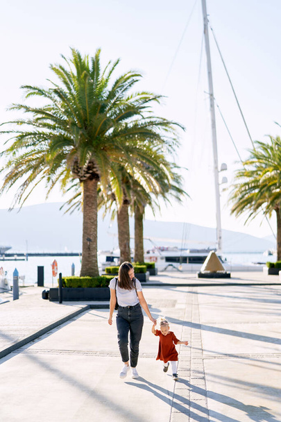 Mother and daughter are running hand in hand among palm trees on a boat pier in Porto Montenegro - Photo, Image