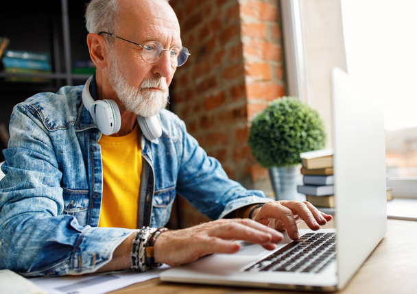 Пожилой мужчина фрилансер в модной одежде, печатающий на клавиатуре ноутбука, сидя за столом и работая в домашнем офисе - Фото, изображение