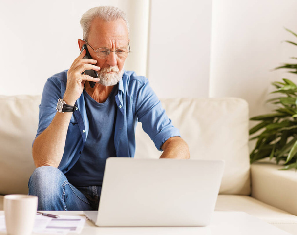 Серьезный сосредоточенный бородатый пожилой человек разговаривает на смартфоне, сидя на диване и читая данные от lapto - Фото, изображение