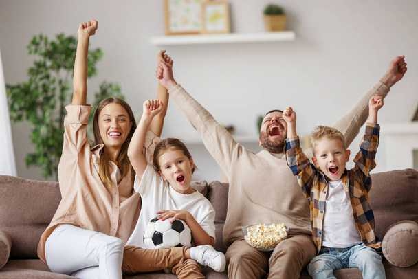 Encantados padres e hijos con palomitas de maíz y bolas levantando los brazos y gritando mientras ven el partido de fútbol y celebran el gol en hom - Foto, imagen