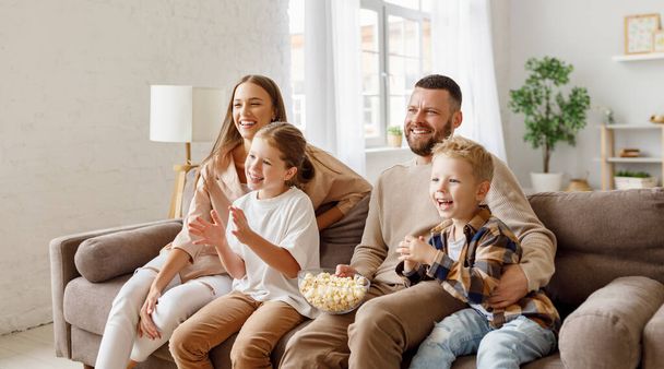 Entzückte Eltern und Kinder, die Popcorn essen und lachen, während sie auf dem Sofa sitzen und lustige Filme bei hom ansehen - Foto, Bild