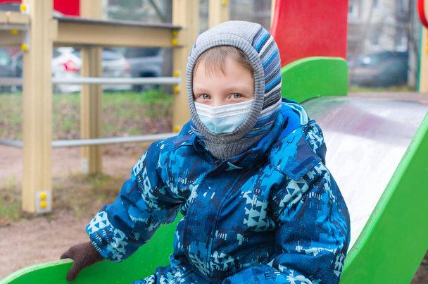 In the fall, a preschooler in a medical mask, alone on the playground,prepares to go down the hill.Concept - children in quarantine, coronavirus pandemic,isolation and social distancing with covid 19. - Zdjęcie, obraz