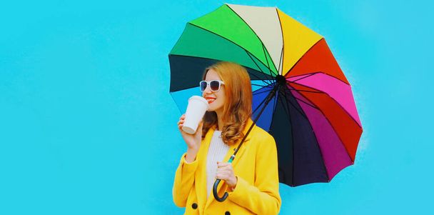 Portrait of young woman drinking coffee with holding colorful umbrella over blue background - Foto, Imagen