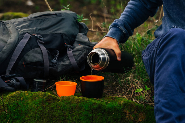 Teetrinken während der Wanderung. Mann gießt heißen Tee aus schwarzer Thermosflasche in 2 Tassen im Wald. Erfrischung beim Wandern. Campingausrüstung - Foto, Bild